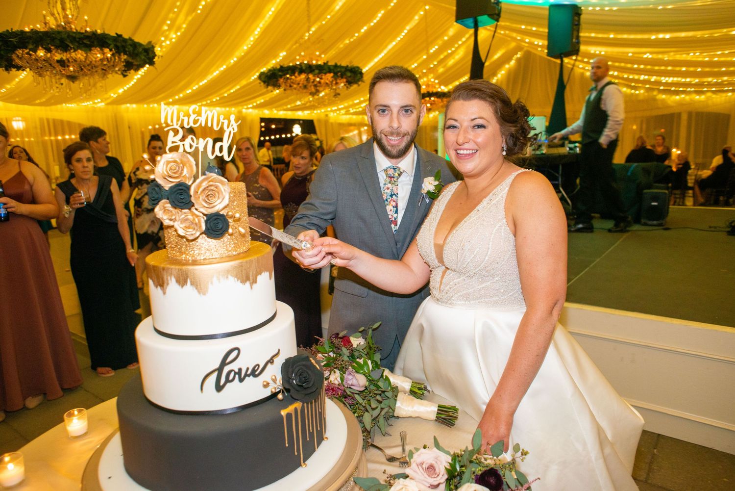 Newlyweds stand beside their elegant black and gold wedding cake at their reception.