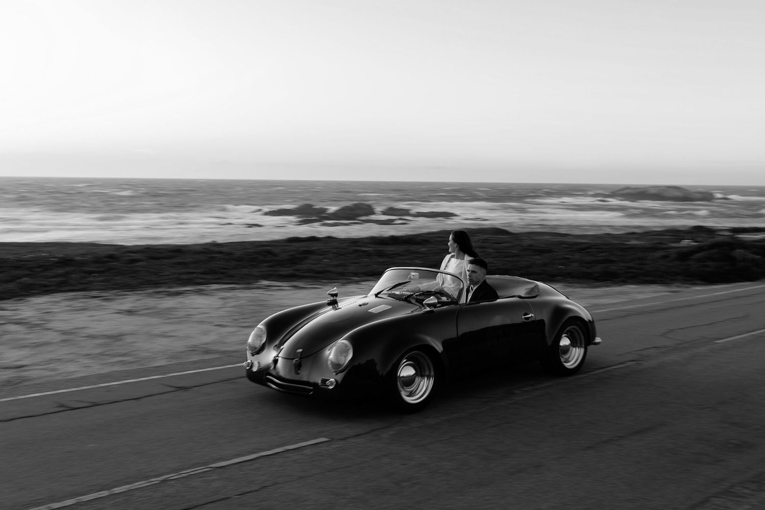 Vintage black convertible Porsche drives along a coastal road in black and white photography.