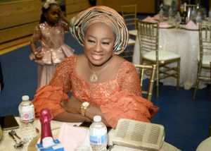 Guest in orange lace dress and traditional headwrap seated at event table with water bottles.