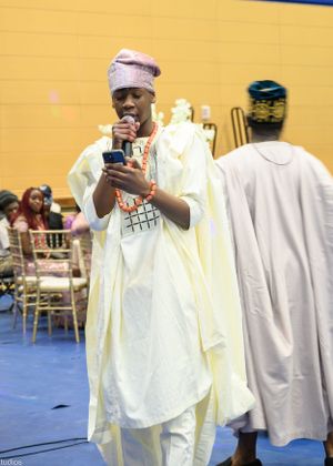 Speaker in traditional white agbada and cap addresses audience at cultural event.