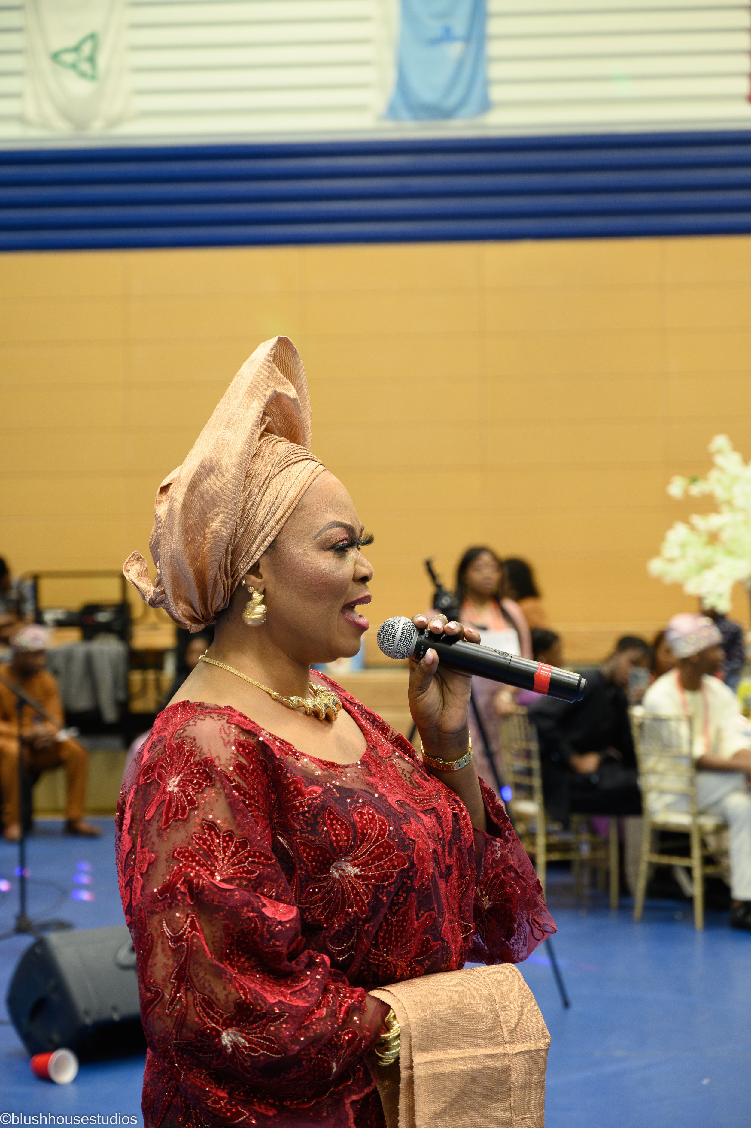 A performer in a red lace dress and peach headwrap sings into a microphone at a special event.