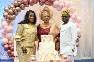 Three guests posing with decorative white cake under balloon arch at celebration.