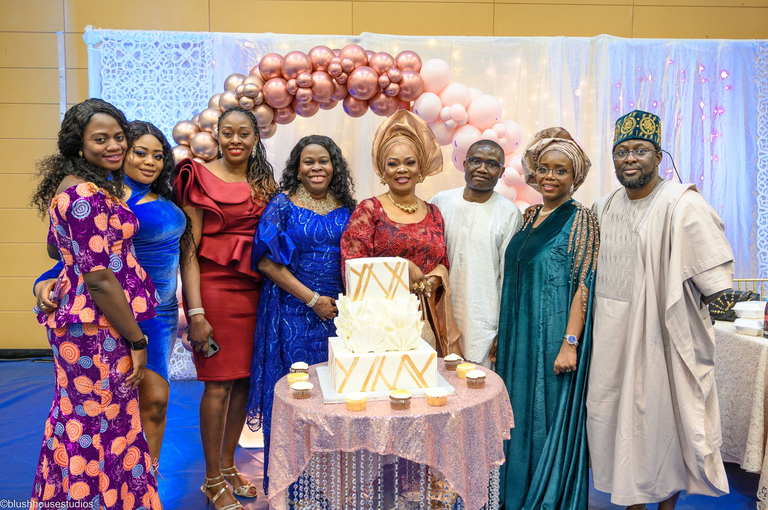 A festive celebration with guests gathered around a white tiered cake under a balloon arch.