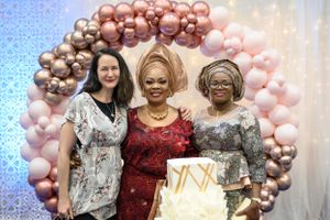 Three guests posing with decorative cake under pink and gold balloon arch at formal event.