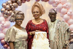 Three guests pose with a white tiered cake under a festive balloon arch.