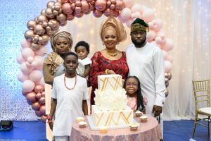Family members gather around a white tiered cake under a rose gold balloon arch.
