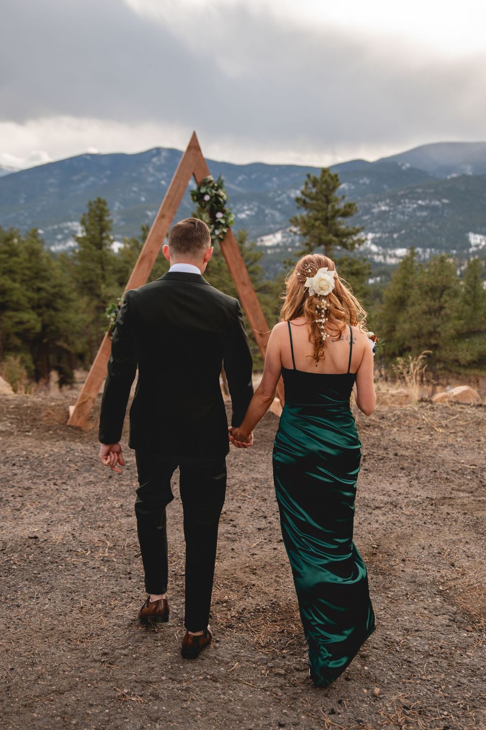 Couple walks toward triangular ceremony arch in mountain setting with emerald dress and black suit.