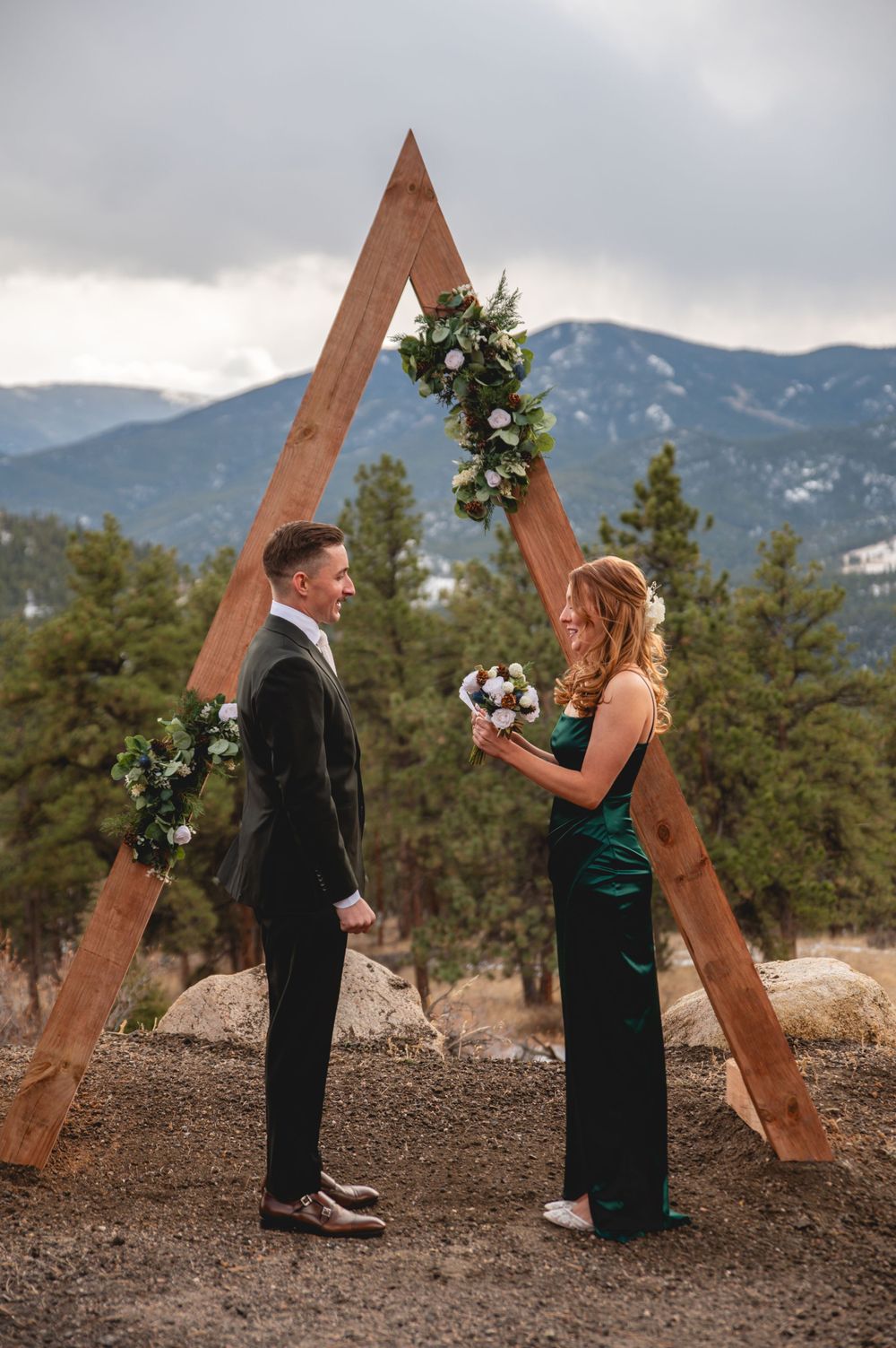 A romantic mountain ceremony under a wooden triangle arch decorated with white florals and greenery.
