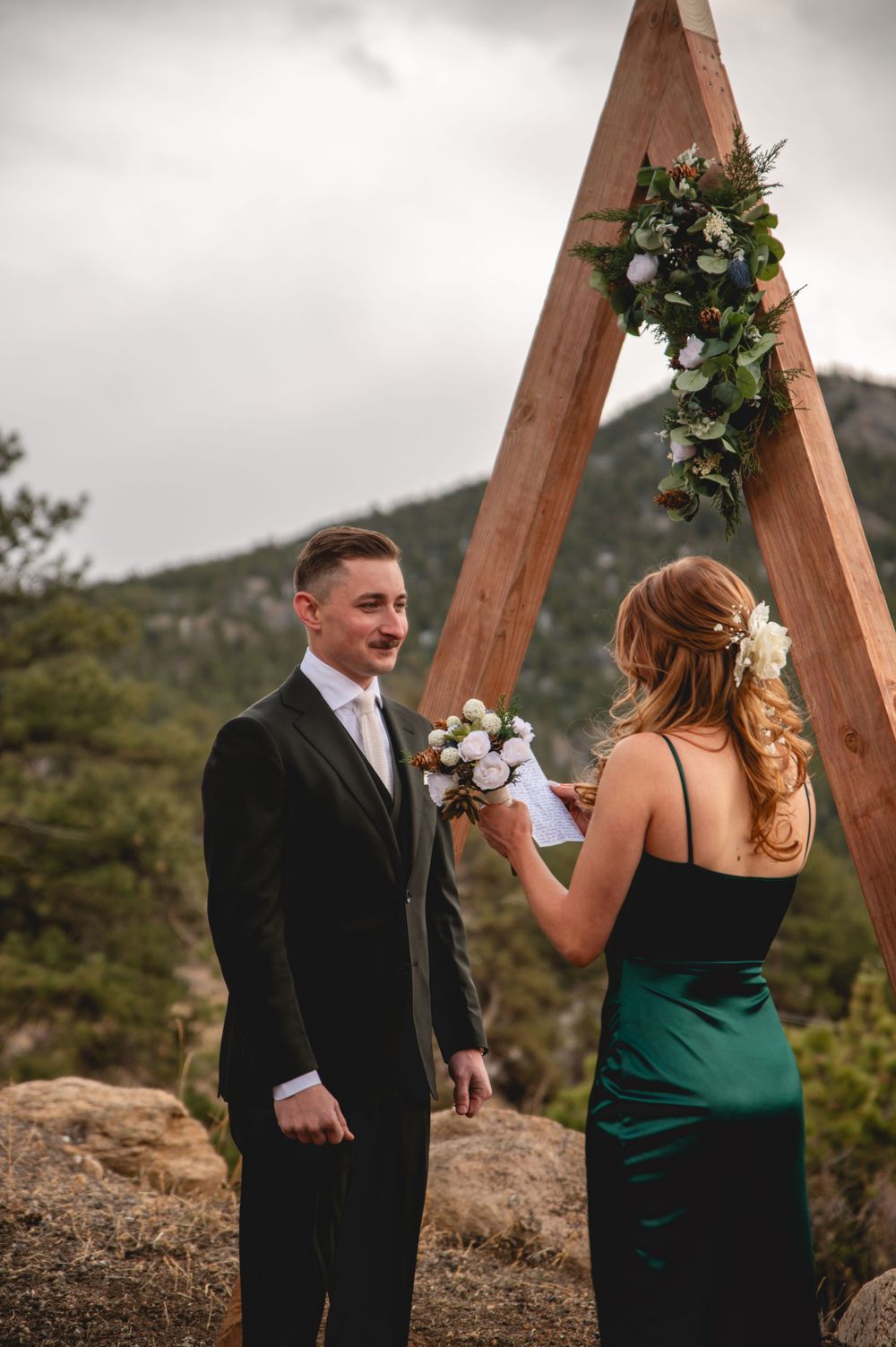Couple shares intimate moment beneath triangular wooden arch with mountain backdrop.
