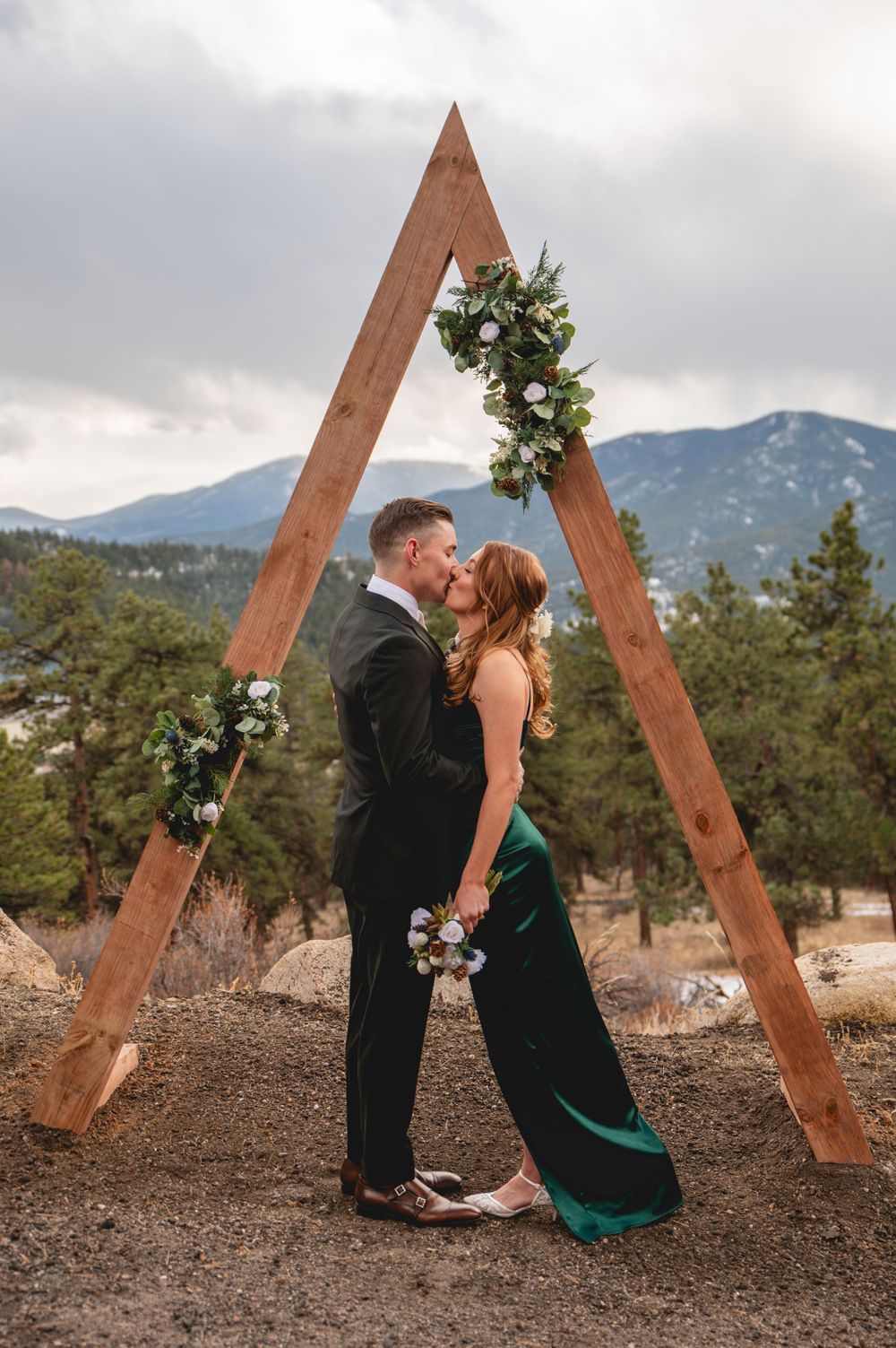 A romantic couple embraces under a wooden A-frame arch decorated with greenery in a mountain setting.