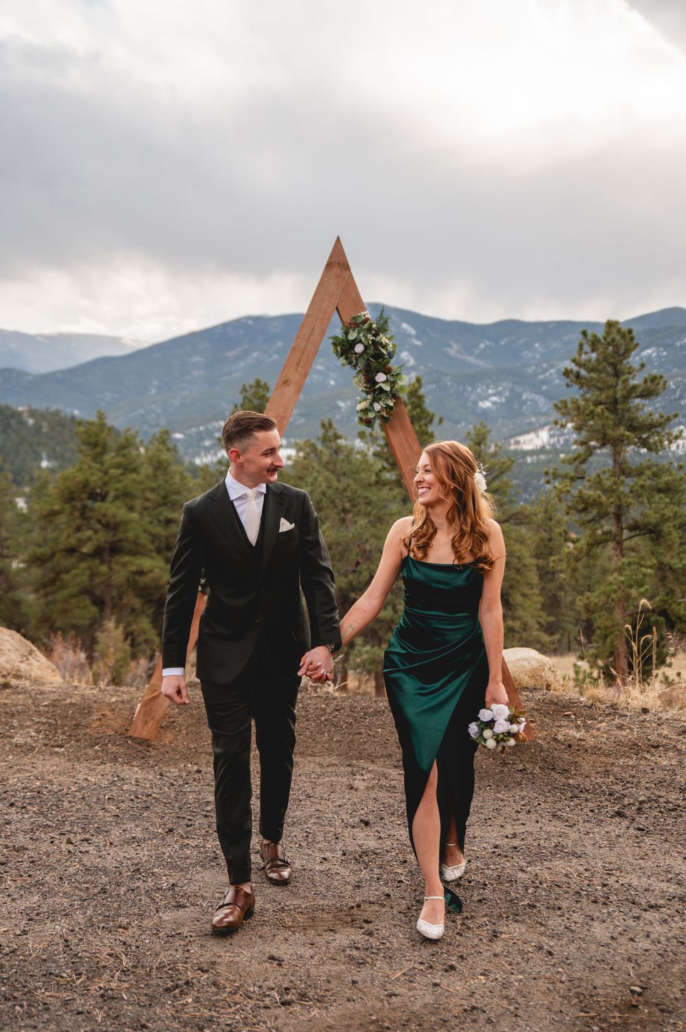 A couple walks hand in hand through a mountain landscape wearing formal attire.