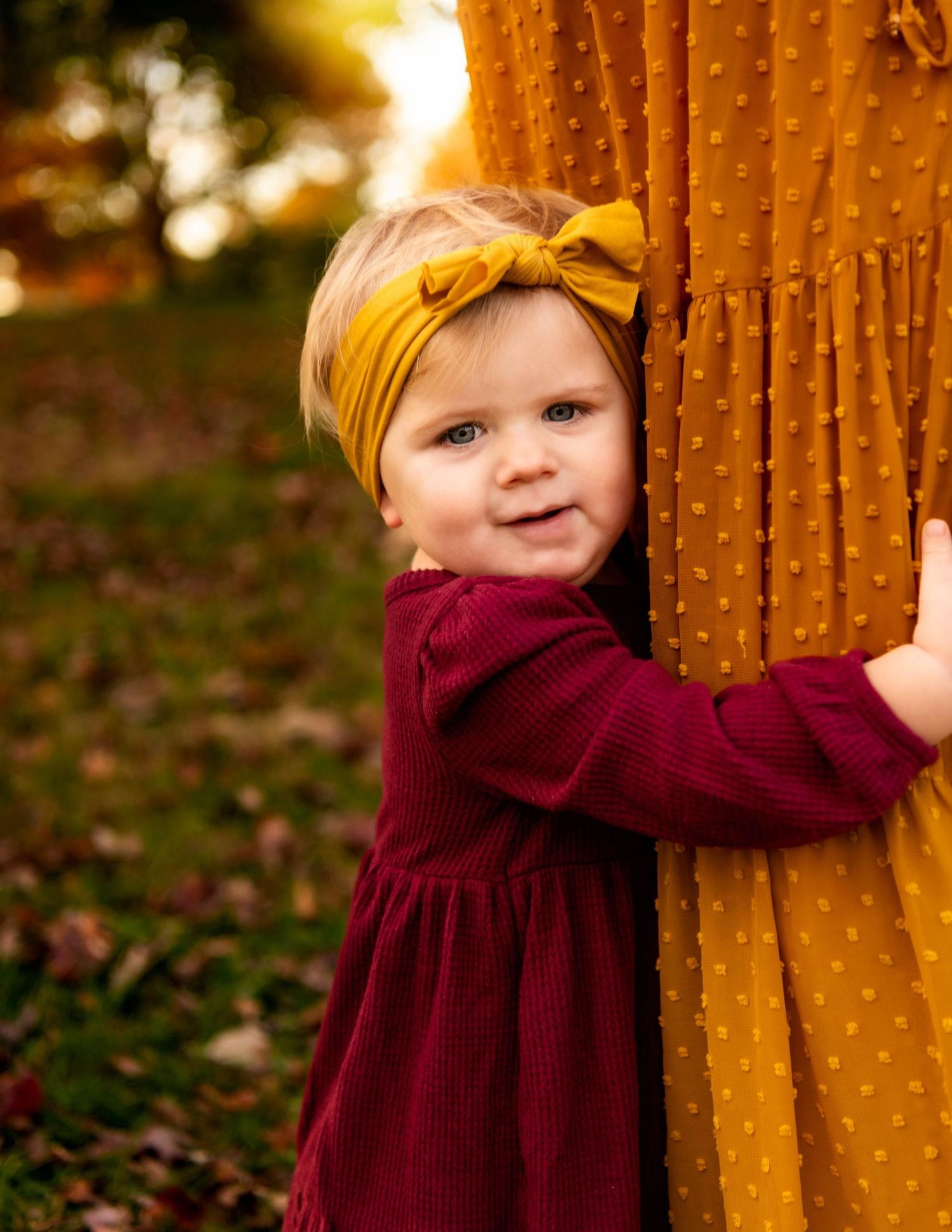 Adorable blonde toddler girl in burgundy dress with bow headband hugs mustard yellow textured fabric outdoors.