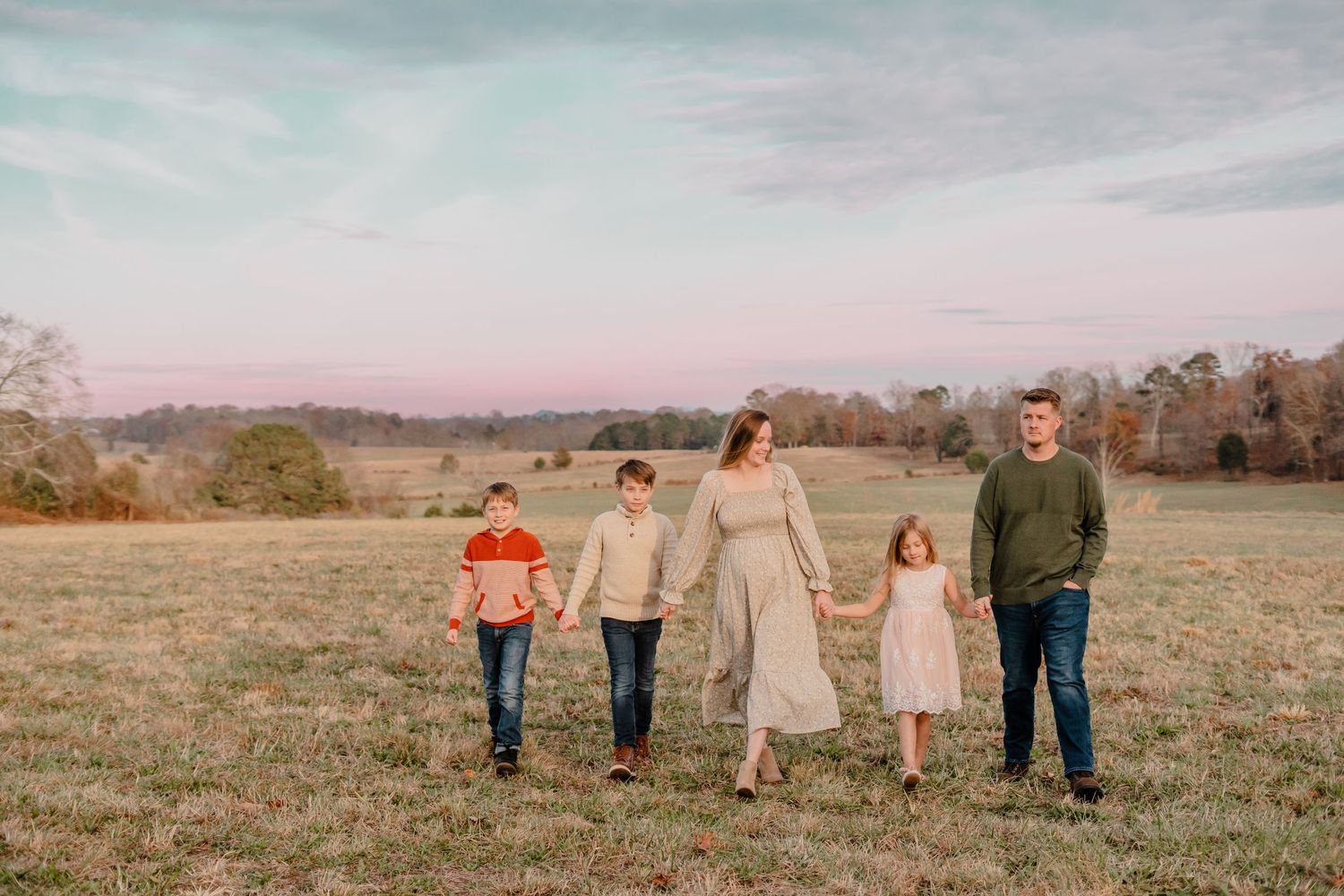Family of five holding hands while walking through an open field at sunset.