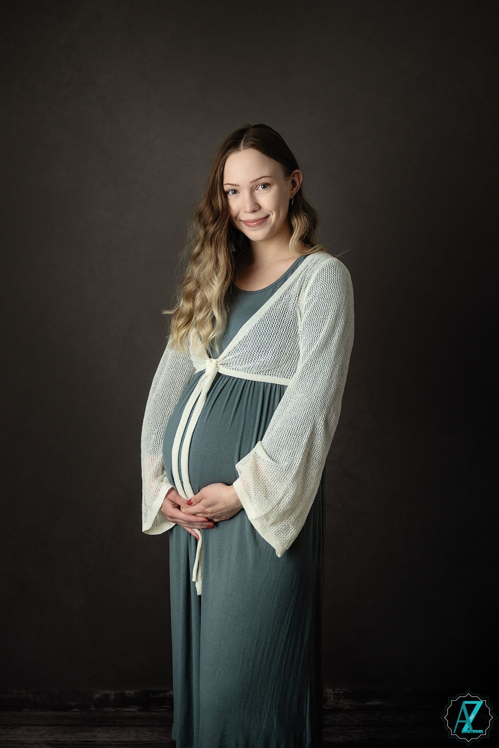 A maternity photo featuring a sage green dress with white cardigan against a dark studio backdrop.
