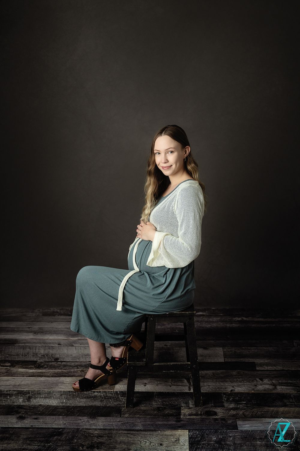 Maternity portrait on wooden floor with subject seated on dark stool wearing flowing green dress.