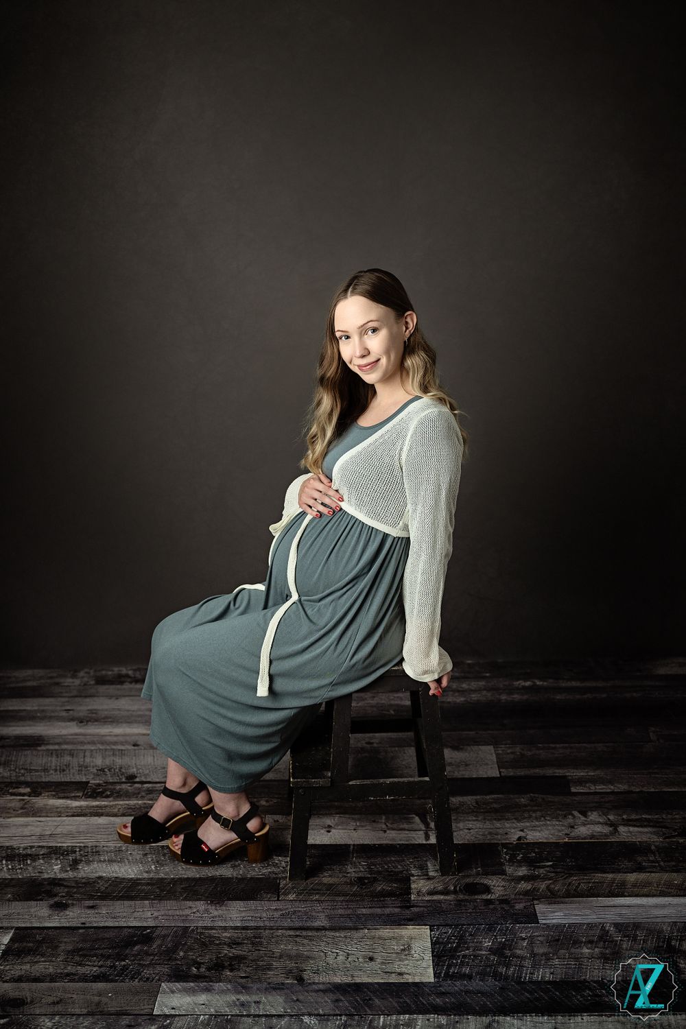 Studio maternity portrait on wooden floor with subject seated wearing sage dress and white sweater.