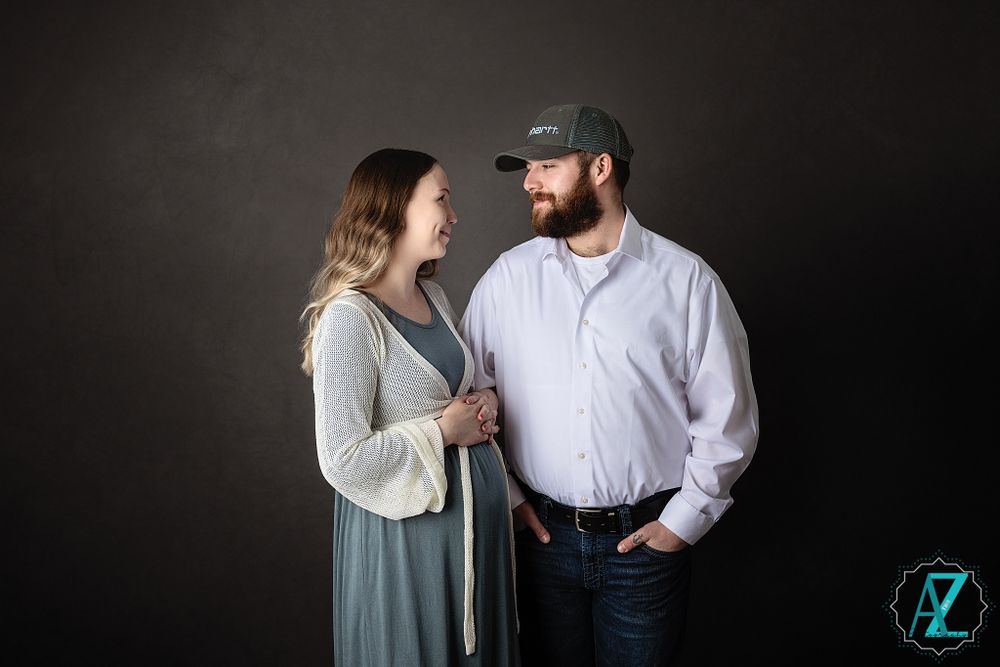 Couple stands together during maternity photoshoot against dark background.