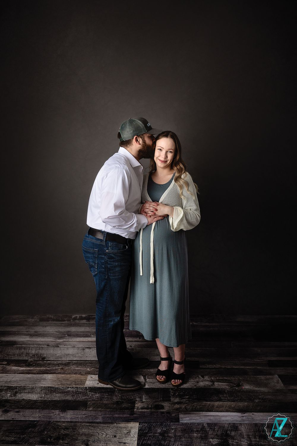 Intimate maternity moment captured against dark background on rustic wooden floor.
