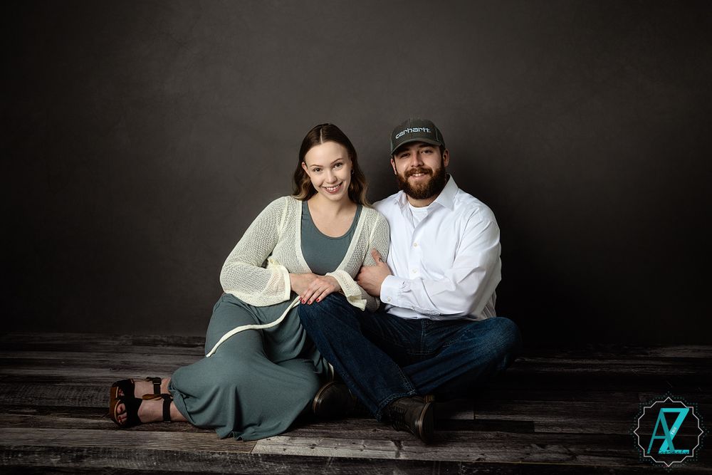 A couple sits intimately on a wooden floor against a dark backdrop for a portrait session.