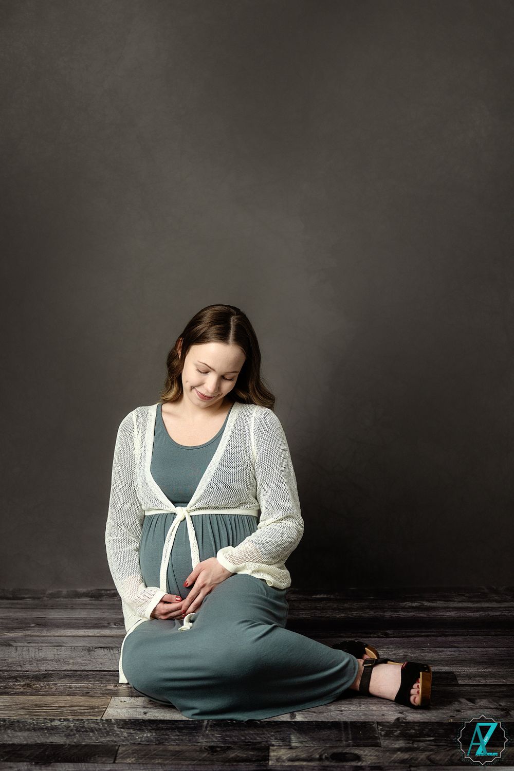 A maternity portrait showing a woman in sage dress and white cardigan sitting on wooden floor.