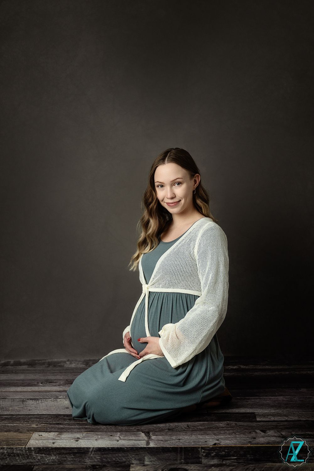 Studio maternity photo of woman in sage dress and white sweater sitting on wooden floor.