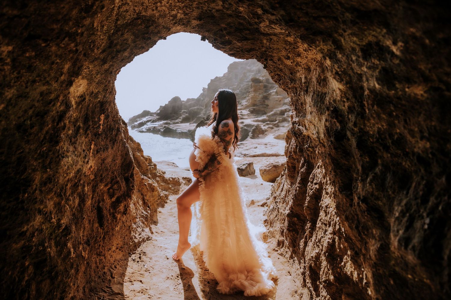 Person in flowing dress photographs ocean view through cave opening at sunset.