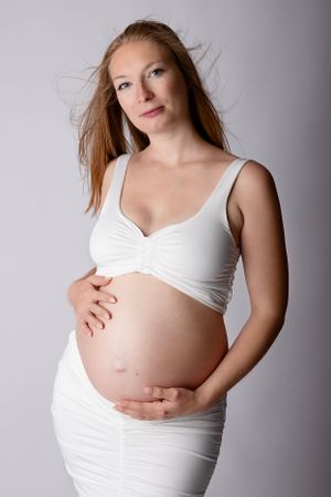 Elegant maternity portrait with expectant mother in white dress against grey background.