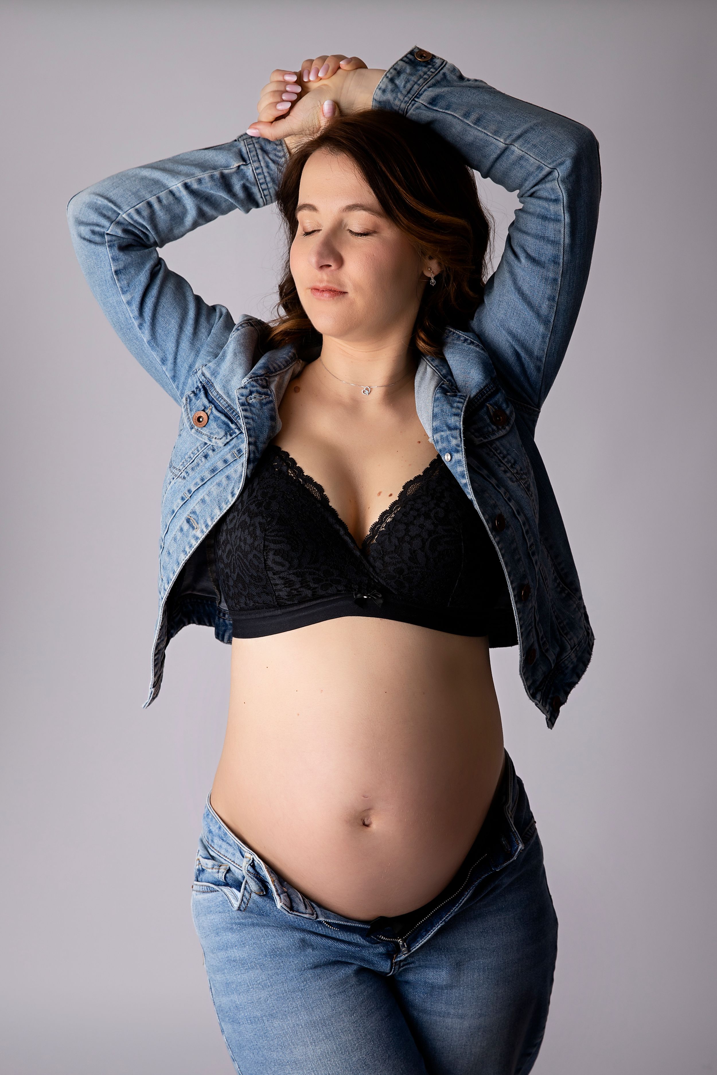 Maternity portrait of woman in denim jacket and jeans showing pregnant belly against grey background.