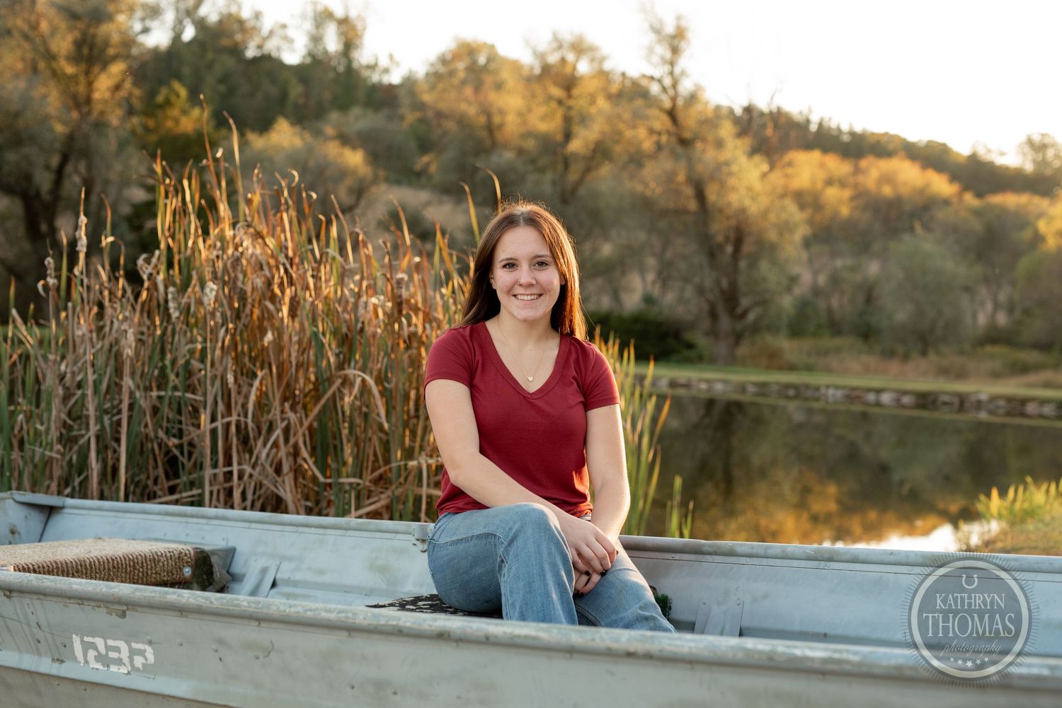 Someone sits on a metal boat by a pond with autumn foliage in the background.