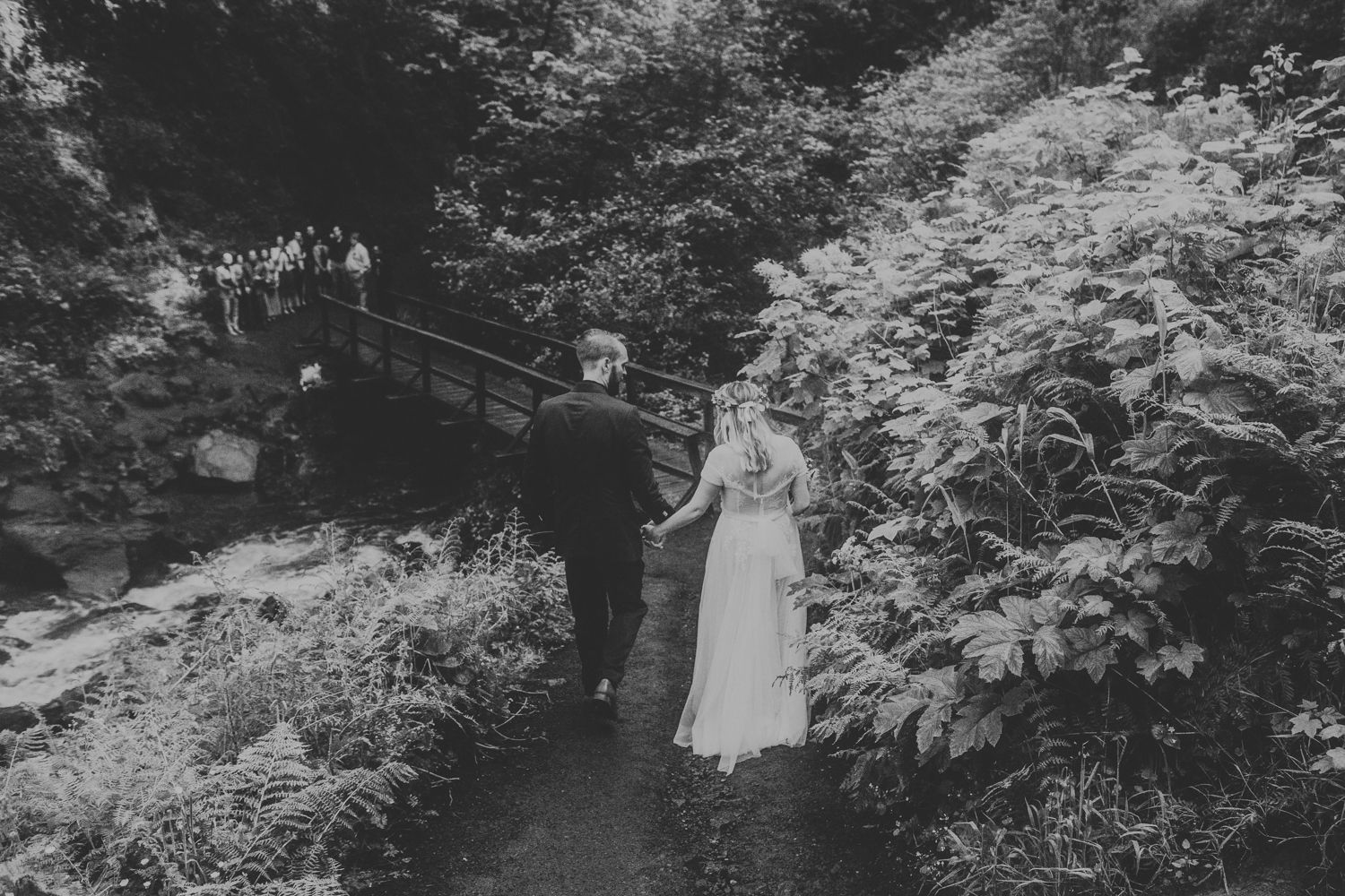A couple walks along a wooded path surrounded by lush green ferns in black and white.