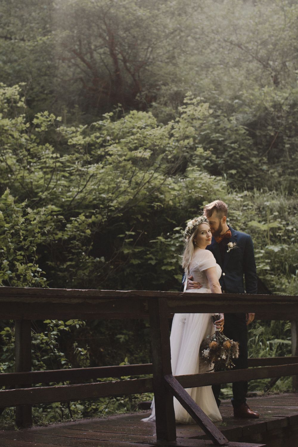 A couple shares a tender moment on a wooden bridge surrounded by lush forest greenery.