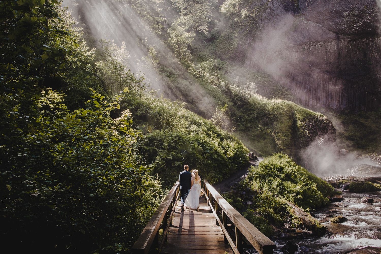 A couple stands on a misty forest bridge with dramatic sunbeams streaming through trees.