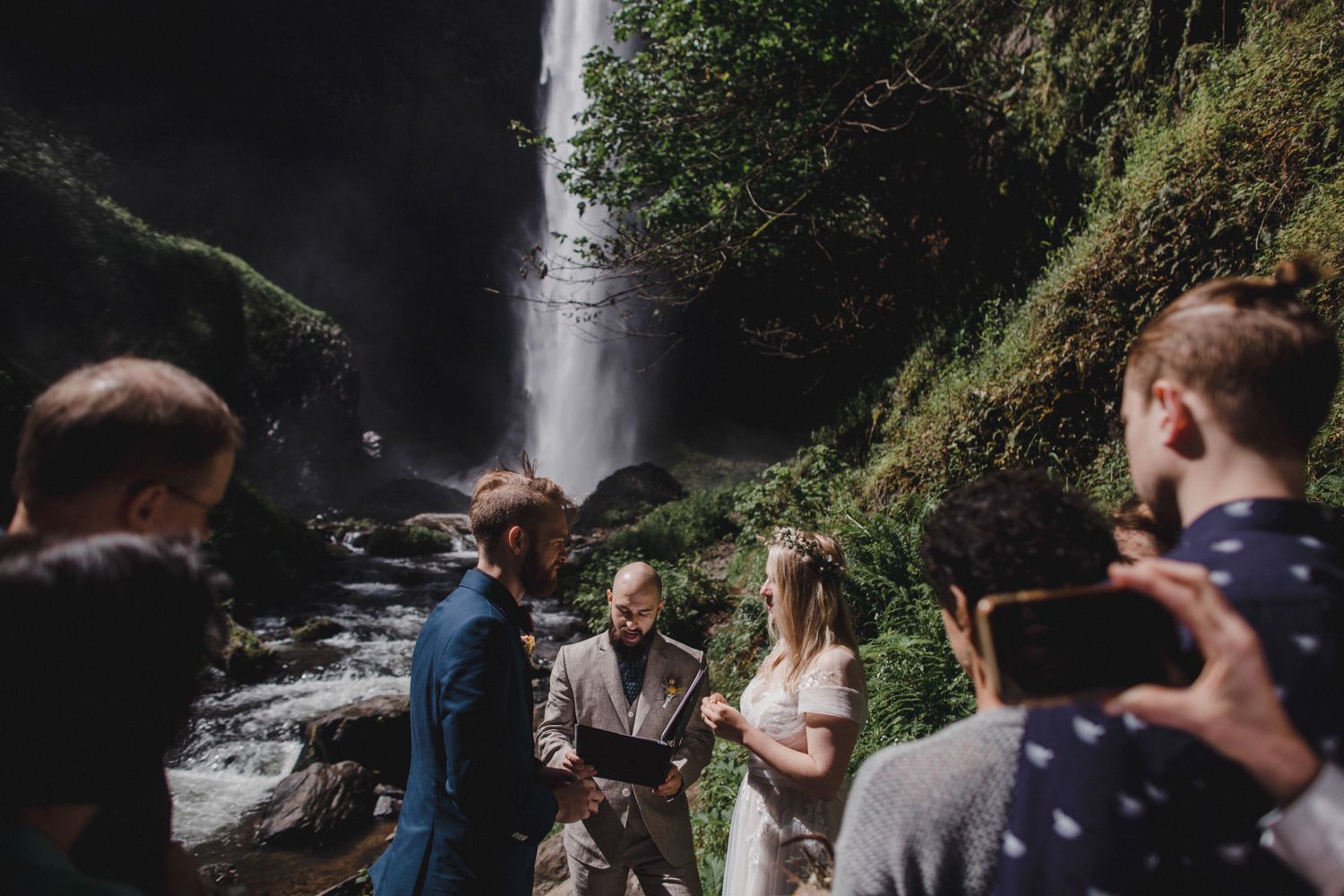 Wedding ceremony taking place in front of a majestic waterfall with guests and officiant gathered on rocky terrain.