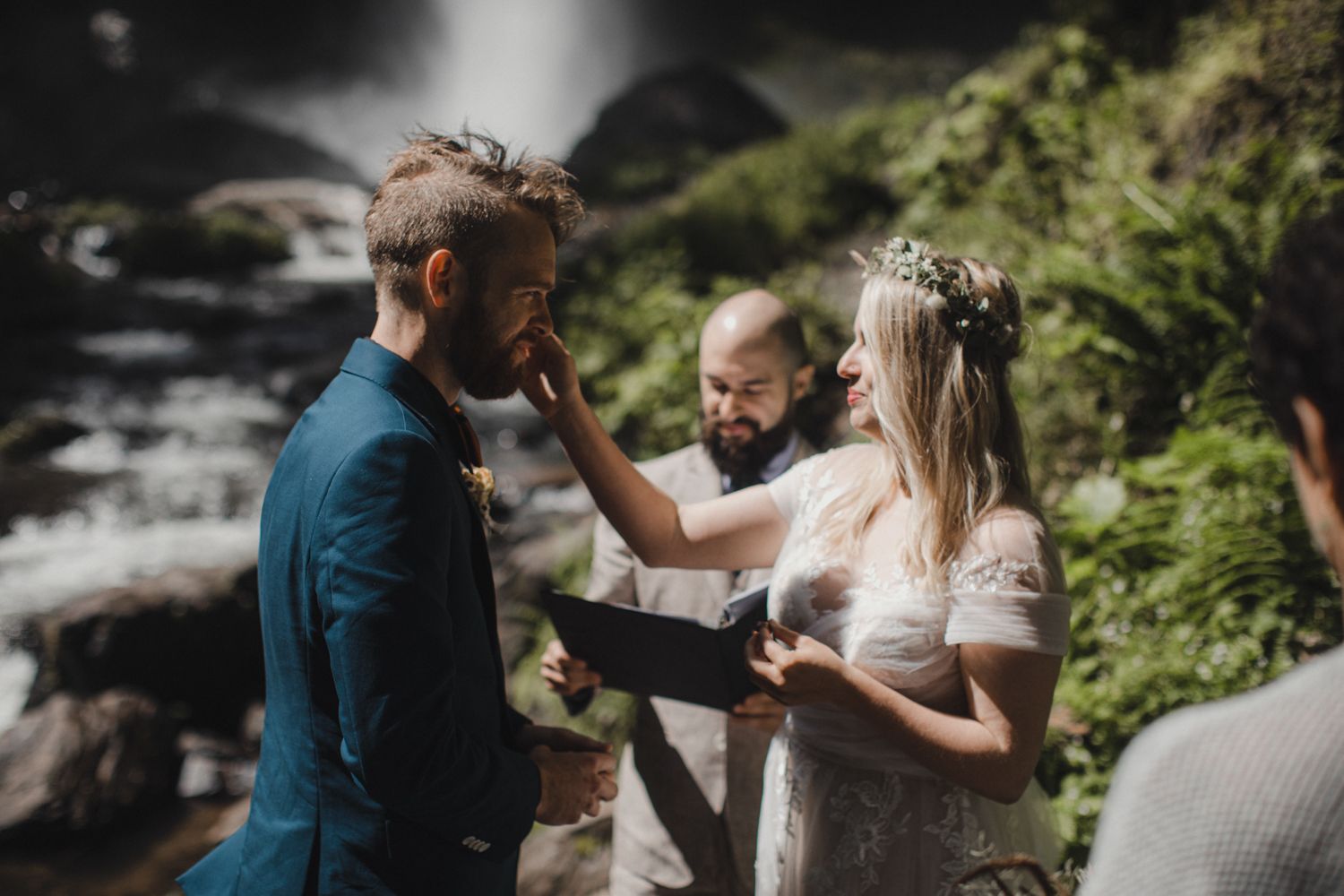 A wedding ceremony takes place by a waterfall with natural light streaming through trees.