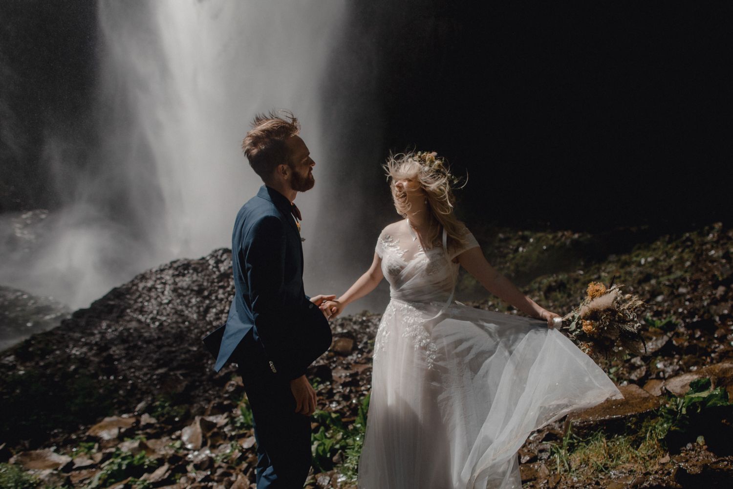 Couple in wedding attire standing together on rocky ground with a dramatic waterfall cascading in the background.