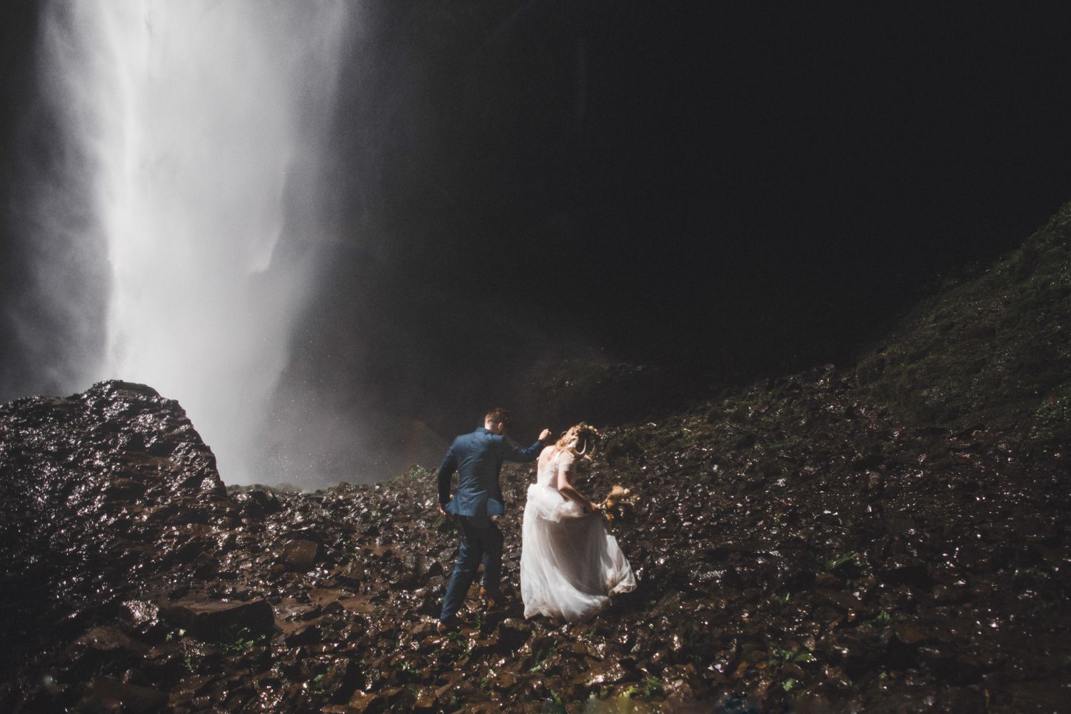Bride and groom walking on rocky terrain beneath a towering waterfall during their outdoor wedding.