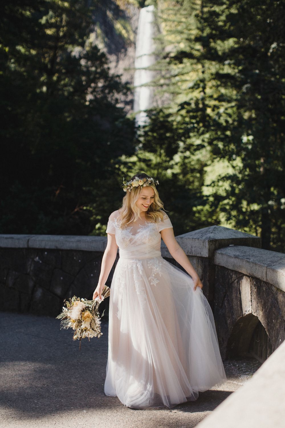 Bride in flowing white dress holding bouquet while standing on stone bridge with waterfall visible behind trees.
