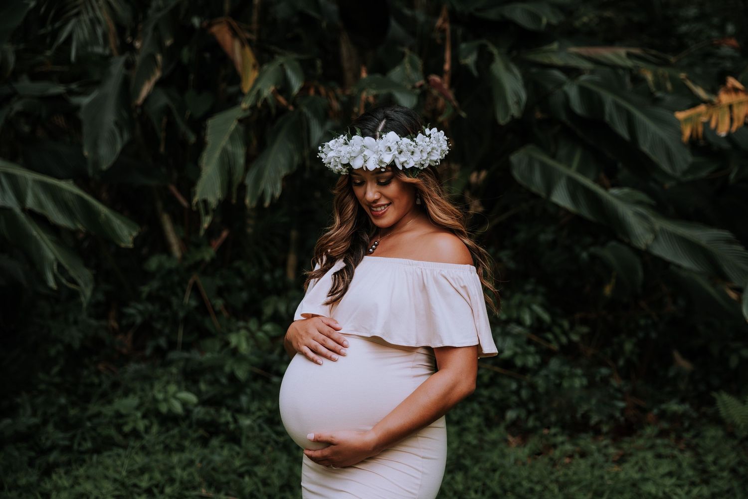 Expectant mother in white off-shoulder dress wearing a floral crown poses against lush greenery in maternity photo.