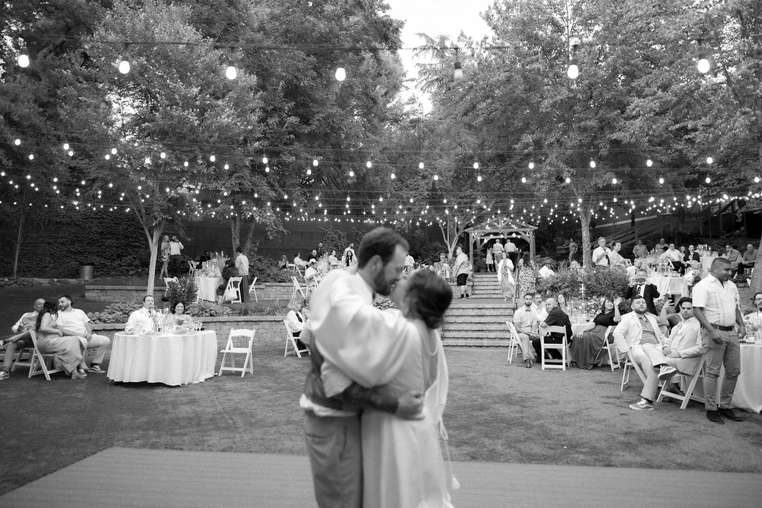 Outdoor evening wedding reception with string lights and couples dancing on lawn.