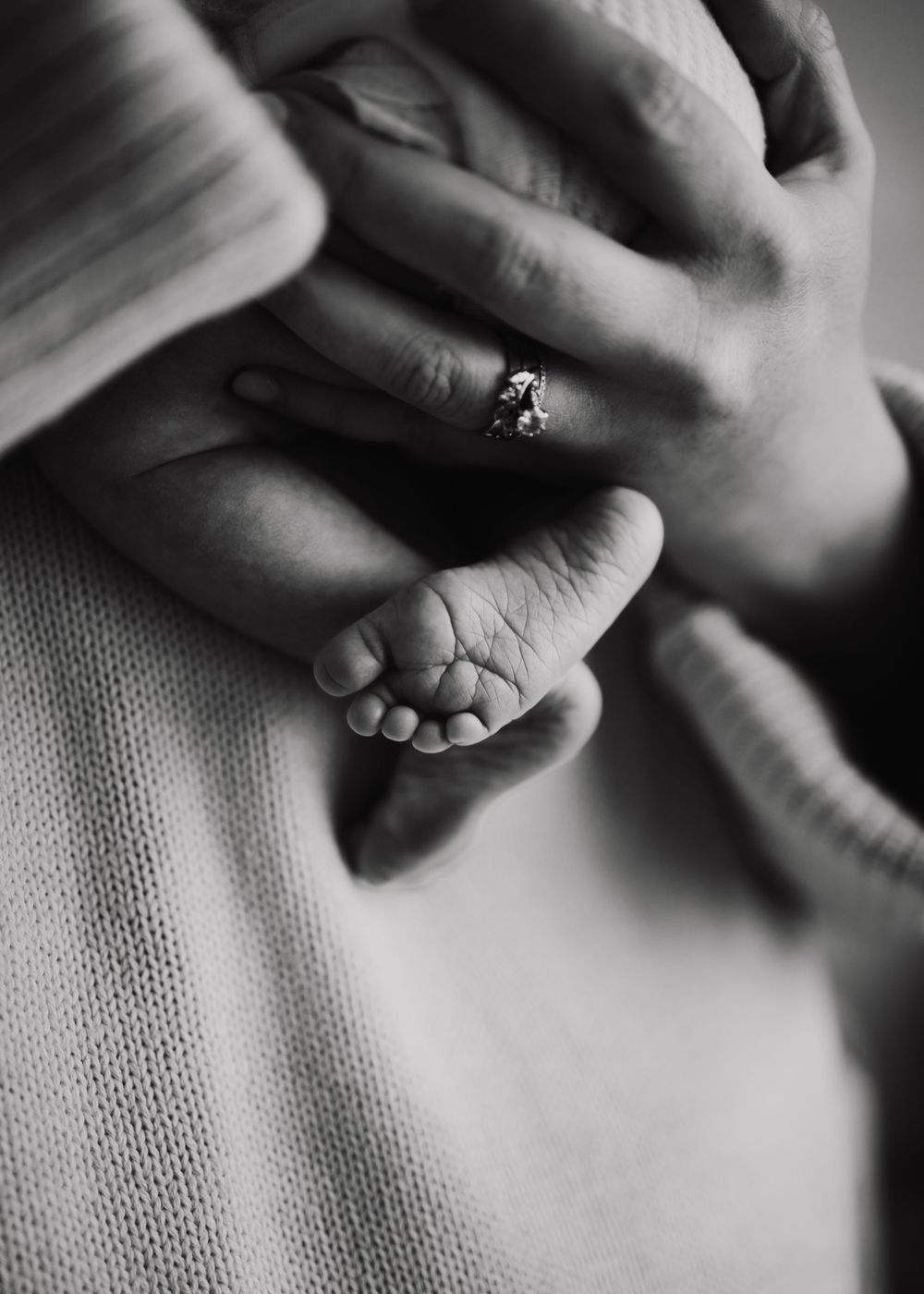 Black and white close-up of tiny baby feet next to adult hands with engagement ring.