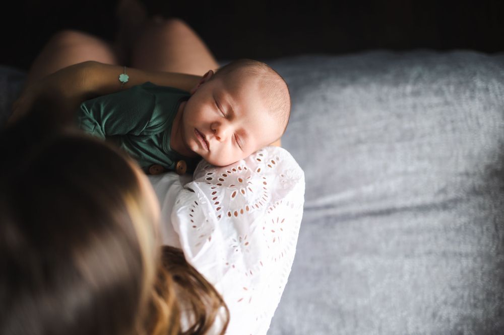A sleeping newborn baby wearing a green outfit rests on white lace fabric in soft window light.