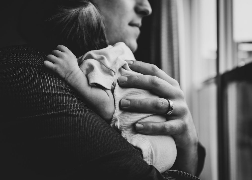 A black and white image showing hands cradling a newborn against a window.