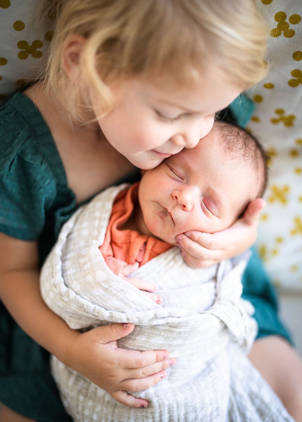 Tender moment between siblings as older child cuddles newborn baby on floral bedding.