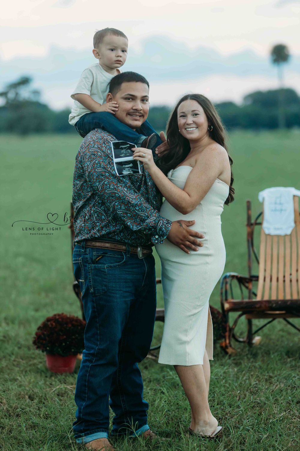 A couple stands together in a grassy field at sunset with a wooden chair in the background.