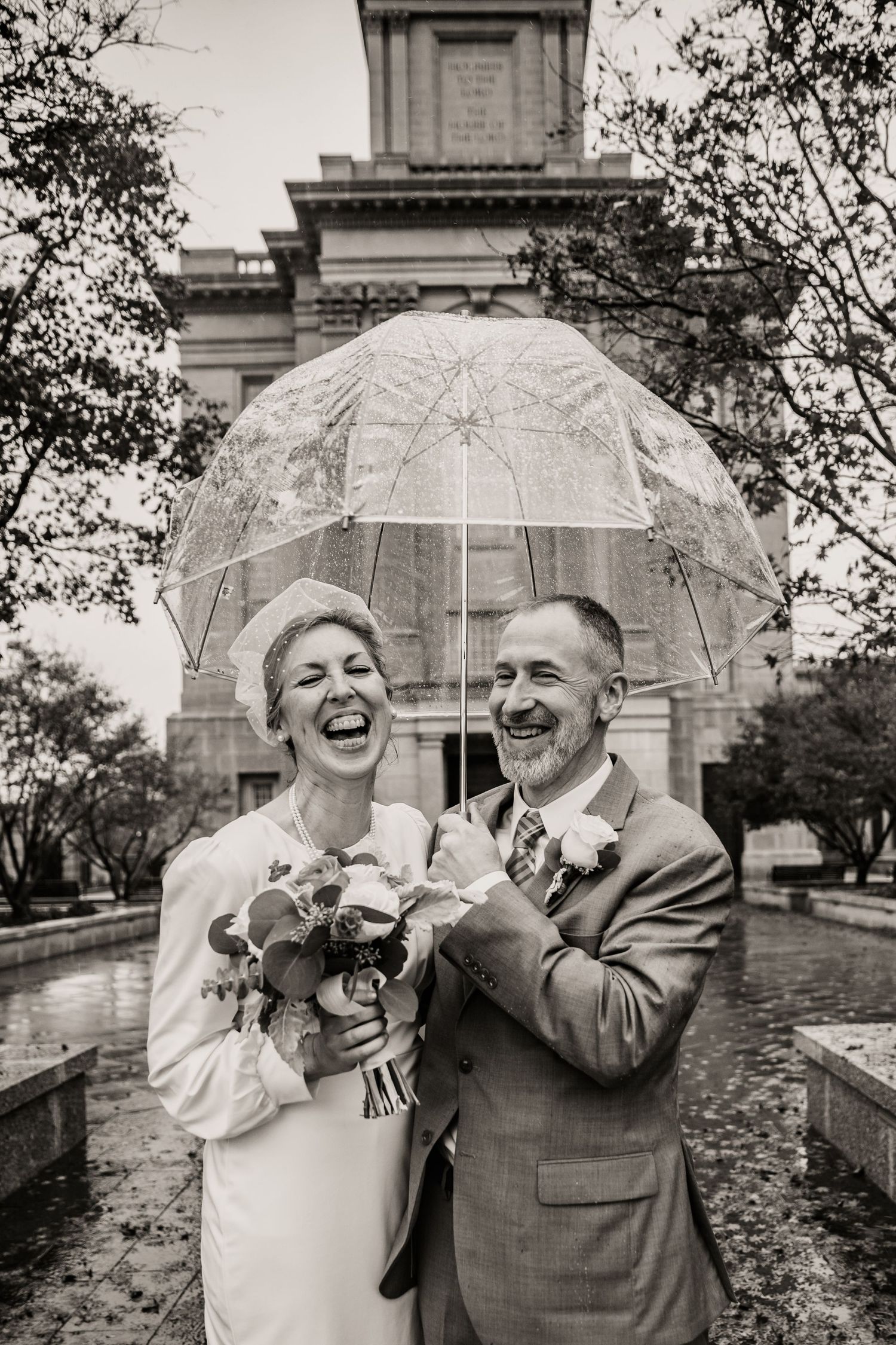 A couple shares a joyful moment under a clear umbrella on a rainy day near historic architecture.