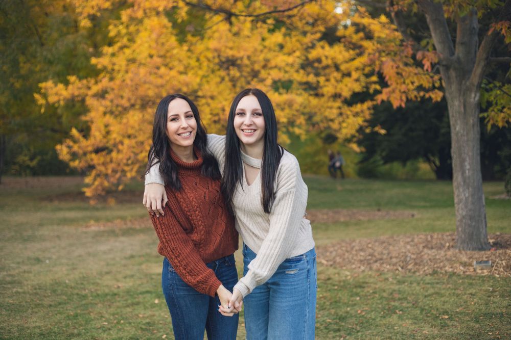 Two friends wearing sweaters stand close together on grass with golden autumn foliage in the background.