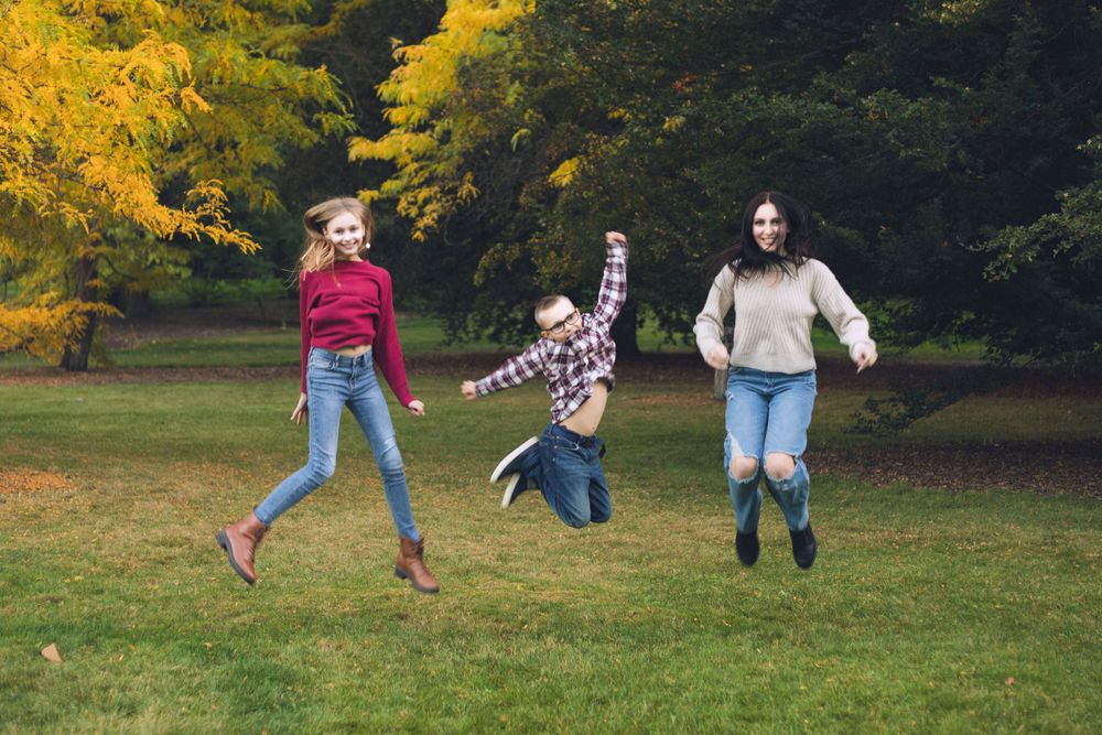 Three people jumping with joy on grass with vibrant fall trees in the background.