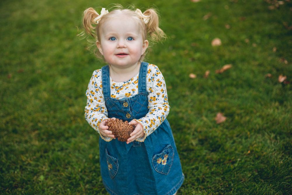 A small child in blue overalls and floral top stands on grass with fallen leaves.