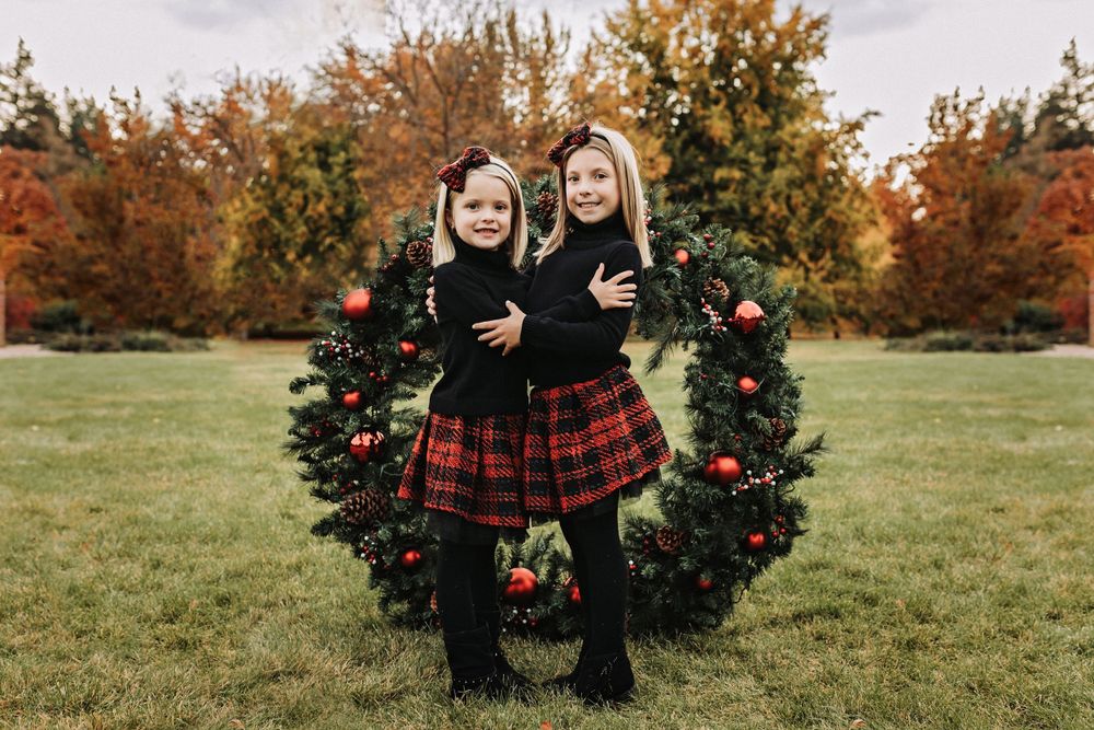 Two matching outfits with plaid skirts pose by a decorated Christmas wreath outdoors during autumn.