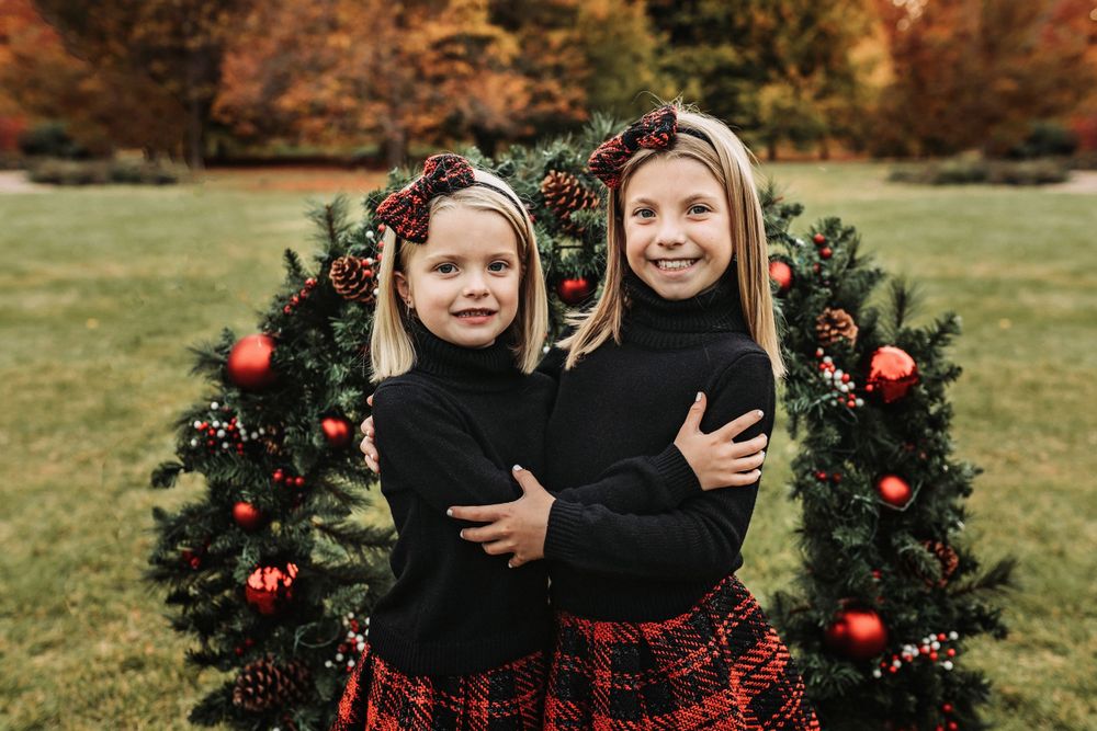 Coordinated black sweaters and plaid skirts pose with Christmas wreath in fall setting.
